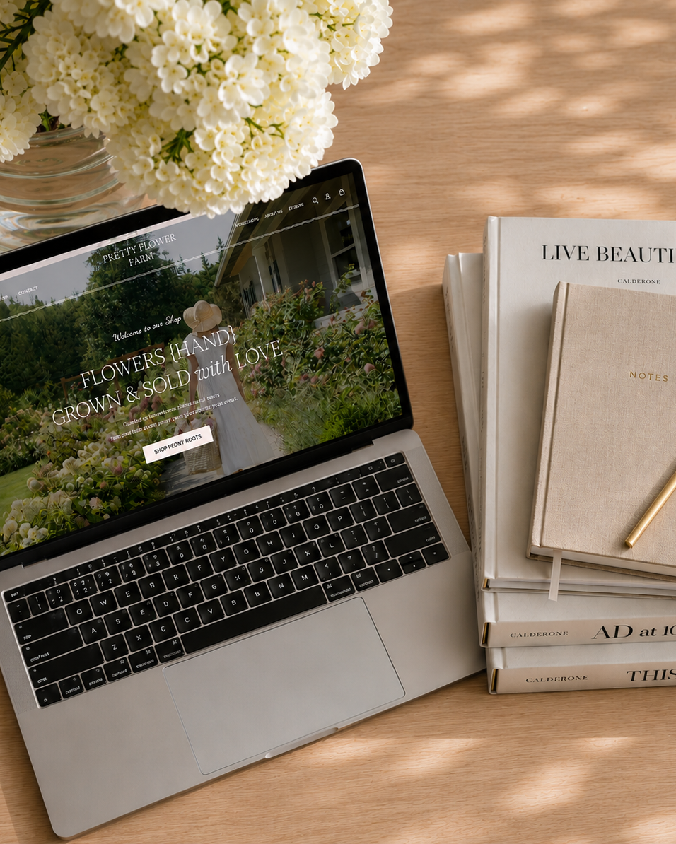 Laptop displaying Pretty Flower Farm's website on a wooden table with books and flowers.